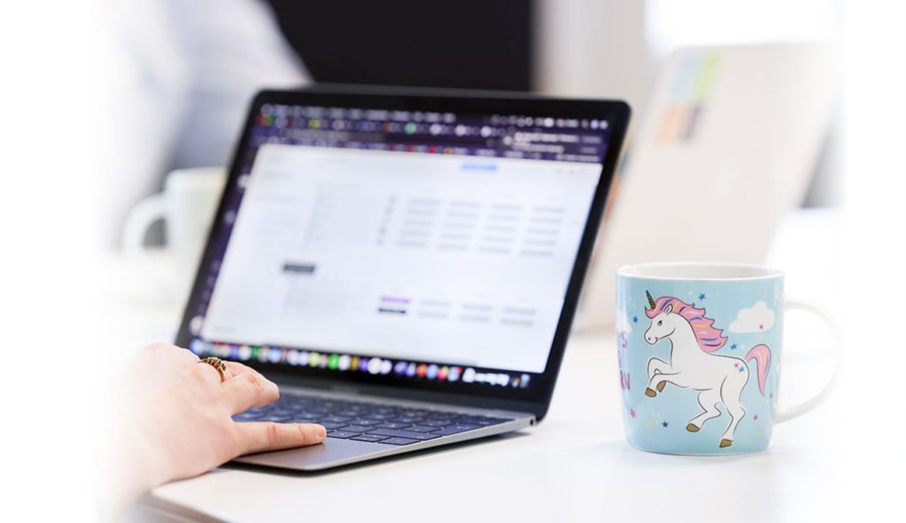 123Consulting Tasse in blau mit weißem Einhorn mit pinker Mähne neben einer Person, welche auf dem Notebook tippt.