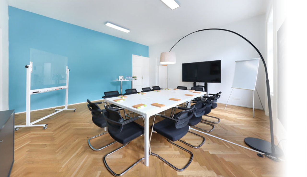 Meetingraum 123 Consulting mittig im Raum langer weißer Tisch, blaue Wand vor Zeichenboard, großer TV und Flipchart